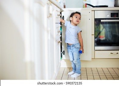 Full-length portrait of playful toddler boy with toy car in hand trying to find something in kitchen cupboard and looking away