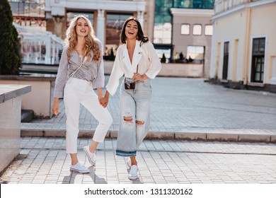 Full-length portrait of pleasant female model in vintage pants holding hands with friend. Outdoor shot of debonair stylish girls standing on the street.
