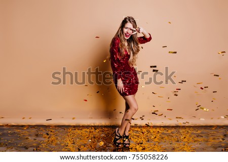 Full-length portrait of pretty dancing girl in trendy black shoes. Long-haired young female model in red sparkle attire fooling around in room with confetti.