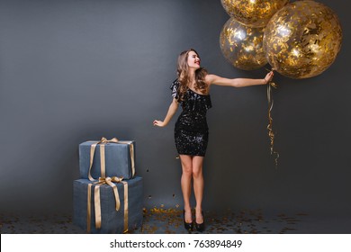 Full-length Portrait Of Refined European Girl Wears Black Dress At Birthday Party. Blissful Long-haired Lady With Balloons Can't Wait To Open Presents.