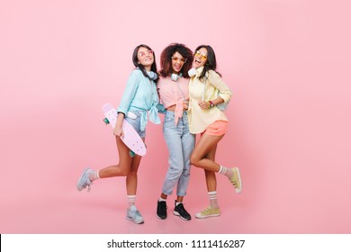 Full-length Portrait Of Skater Girl Posing With Best Friends On Pink Background. Indoor Photo Of Three Ladies In Sport Shoes Laughing Together During Joint Photoshoot.