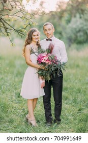 The Full-length Portrait Of The Smiling Bridesmaid In The Knee Length Dress Holding The Huge Bouquet And The Smiling Best Man Hugging With Her At The Background Of The Forest.