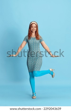 Full-length portrait of smiling, young, redhead woman with stylish makeup and hairdo posing against blue studio background. Concept of retro style, fashion, beauty, elegance, 60s, youth. Pin-up style