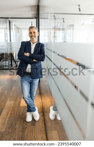 Full-length portrait of a stylish mature businessman standing in the modern office and posing against the glass partition with arms crossed, male employee, manager looking at the camera, vertical