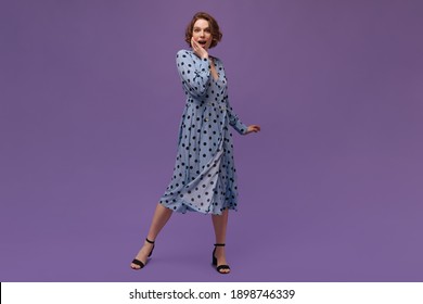 Full-length portrait of trendy girl against violet plain background. Lady in blue polka dot dress and black shoes looking straight forward surprisingly and posing at studio
