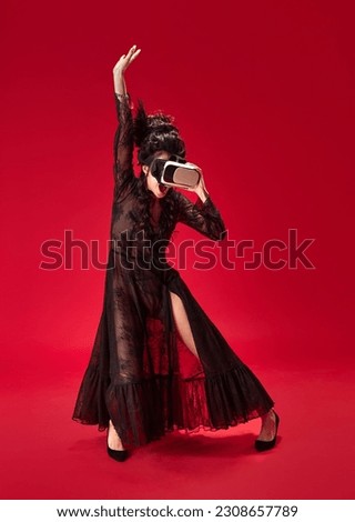 Full-length portrait of young girl in elegant, stylish black dress wearing VR glasses and dancing against red background. Concept of history, renaissance art, comparison of eras, modern technologies