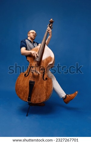 Full-length portrait of young man, musician, emotionally, excessively playing double bass against blue studio background. Concept of music, talent, hobby, entertainment, festival, performance, ad