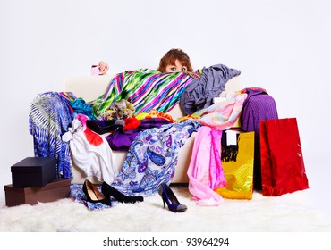 Full-length Portrait Of Young Shopaholic Woman With Credit Cards Hiding Behind Sofa With A Lot Of Clothes And Shopping Bags