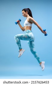 Full-length Portrait Of Young Sportive Girl Training, Jumping With Dumbbells Isolated Over Blue Studio Background. Concept Of Sport, Fitness, Healthy Lifestyle, Strength, Youth, Ad