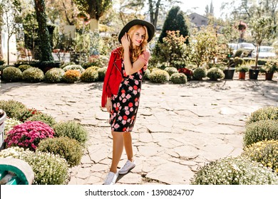 Full-length Shot Of Fair-haired Woman In Black Dress With Floral Print, Stylish Hat And Black Leather Jacket Walking In Garden