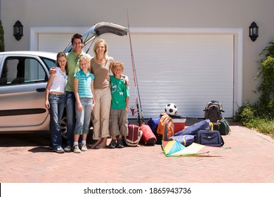 Full-length Shot Of A Family Standing Beside The Parked Car With Open Boot Near Camping Equipment On The Driveway.