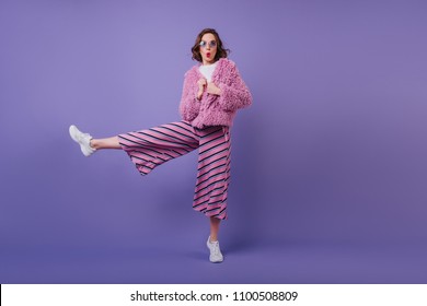 Full-length shot of inspired dark-haired girl standing on one leg. Caucasian woman expressing amazement while posing with kissing face expression on purple background.