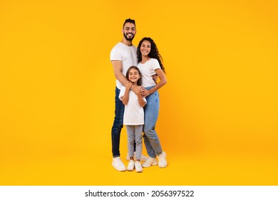 Full-Length Shot Of Middle-Eastern Family On Yellow Background. Happy Arabic Parents Hugging Their Little Daughter Standing Together And Smiling To Camera Posing In Studio