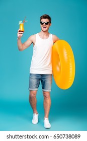 Full-length shot of young brunette man with swimming circle ans cocktail posing at camera isolated over blue