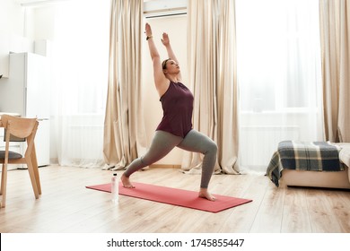 Full-length Shot Of Young Curvy Woman In Sportswear Standing In Warriror I Pose On A Yoga Mat At Home. Determination, Will-power, Sport Concept. Horizontal Shot