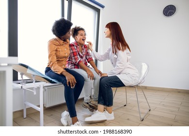 Full-length Shot Of Young Female Laryngologist Doctor Examining Afro-american Child Patient Throat With Tongue Depressor. Pretty Young Black Mother Supporting Her Daughter At Doctor's Office