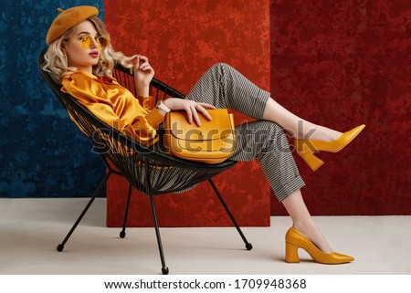 Full-length studio fashion portrait of elegant woman wearing yellow color sunglasses, beret, silk blouse, houndstooth printed trousers, pointed toe shoes, posing on chair, holding stylish leather bag
