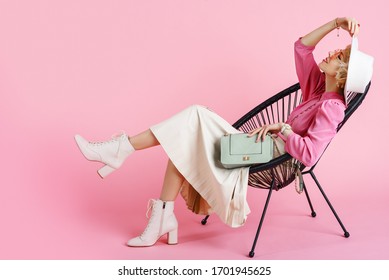 Full-length studio fashion portrait of young elegant happy smiling woman wearing trendy spring outfit: pink blouse, white hat, pleated skirt, ankle boots, holding stylish faux leather bag