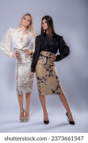 Full-length studio portrait of a two beautiful sexy slim young caucasian brunette and blonde women standing on high heels in designer clothes on white grey background attractively posing