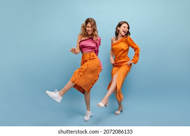 Full-length Two Beautiful Cheerful Caucasian Young Girls Are Dancing In Photo Studio. Blonde In Pink Top, Skirt. Brown-haired Woman In Orange Dress With Bare Shoulder In Shoes.