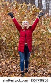 Full-length Vertical Photo Of Pretty Smiling Young Woman With Long Blond Hair Wearing Red Puffy Winter Coat Looking Up While Throwing Dry Leaves Over Her Head