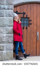 Full-length Vertical Photo Of Pretty Smiling Young Woman With Long Blond Hair Wearing Red Puffy Winter Coat And Jeans Leaning In Stone Porch In Front Of Old Wooden Door