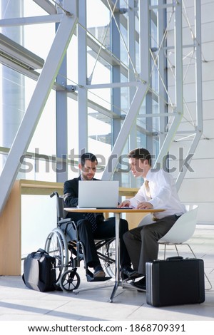 A full-length vertical view of a businessman in wheelchair using laptop with his co-worker in a modern office