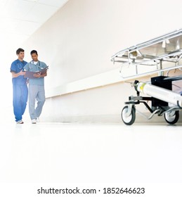 A Full-length Wide Shot Of A Hospital Corridor With Doctors In Scrubs Discussing A Medical Chart