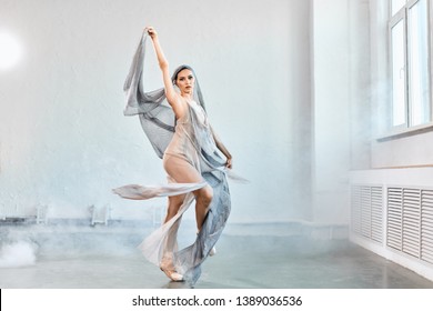 Full-size Expressive Photo Of Female Ballet Dancer Dressed In Grey-blue Flowing Fabric Spinning On One Leg On White Y Background.