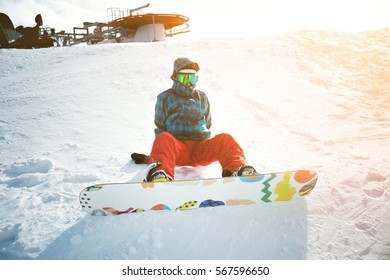Fully equipped and covered from cold beginner girl snowboarder wears her google mask, sits alone at top of ski slope near ski lift in sun rays, ready to stand up and ride down