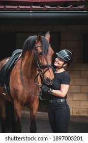 Fully Equipped Female Rider With Helmet And Black Suit In Front Of A Stable With Her Brown Horse Looking Into Her Eyes And Smiling