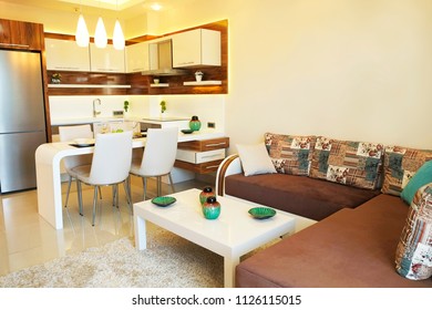 Fully stacked open plan kitchen living room, minimal modern design interior apartment. White furniture, upholstery. Counter w tableware, table settings, oven, sofa, shelves, decor elements. Background