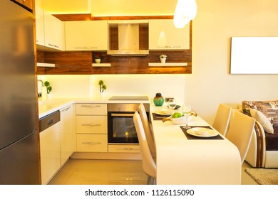 Fully stacked open plan kitchen living room, minimal modern design interior apartment. White furniture, upholstery. Counter w tableware, table settings, oven, sofa, shelves, decor elements. Background