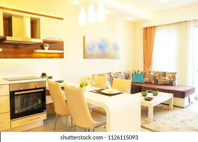 Fully stacked open plan kitchen living room in minimal modern design interior apartment w/ full length windows. Counter w/ tableware, table settings, oven, sofa, shelves, decor elements. Background.