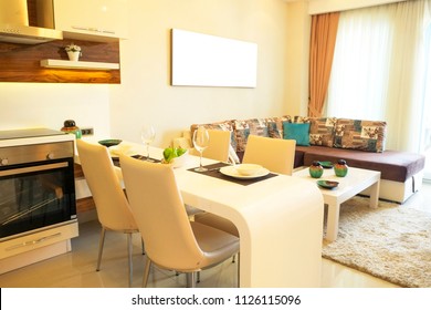 Fully stacked open plan kitchen living room in minimal modern design interior apartment w/ full length windows. Counter w/ tableware, table settings, oven, sofa, shelves, decor elements. Background.