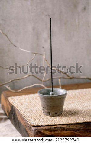 Fuming incense stick in hand crafted gray ceramic incense holder on wooden table with dry branch, grey wall at background.