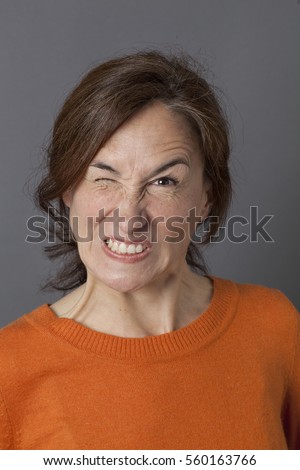 fun angry expression for winking middle aged woman showing her teeth for mother's exasperation and crazy frustration, grey background