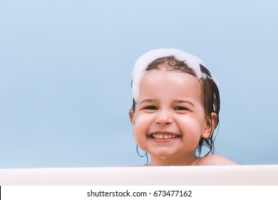 Fun Cheerful Happy Toddler Baby Taking A Bath Playing With Foam Bubbles. Little Child In A Bathtub. Smiling Kid In Bathroom On Blue Background. Infant Washing And Bathing. Hygiene And Health Care.