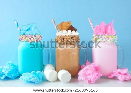 Fun colorful summer milkshakes in mason jar glasses against a pastel blue background. Blue and pink cotton candy and chocolate smores sweet drinks.
