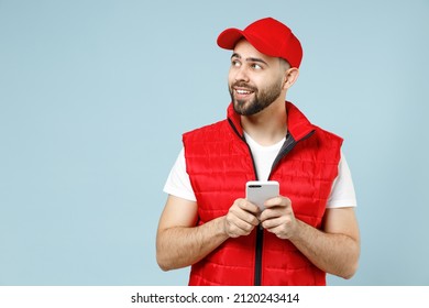 Fun delivery guy employee man in red cap white T-shirt vest uniform workwear work as dealer courier hold use mobile cell phone isolated on pastel blue color background studio portrait. Service concept