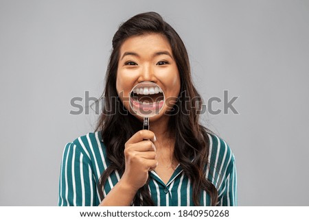 fun, dental care and people concept - happy asian woman showing her teeth through magnifying glass over grey background