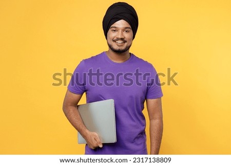 Fun devotee Sikh Indian IT man ties his traditional turban dastar wear purple t-shirt hold use work on laptop pc computer isolated on plain yellow background studio portrait. People lifestyle concept