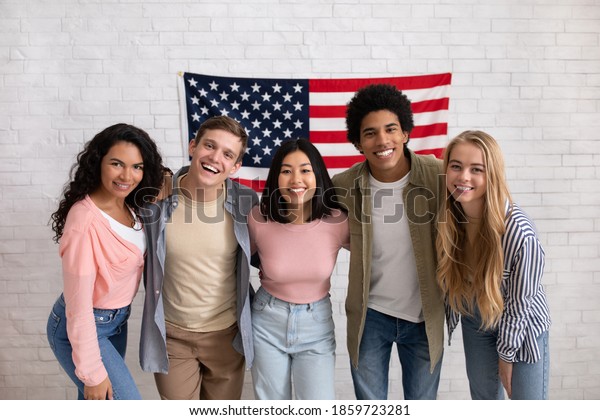 Fun education, language courses, learn english and\
exchange students. Young smiling people of different nationalities\
hug in college with large USA flag on white brick wall, free space,\
studio shot