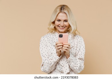Fun elderly gray-haired blonde woman lady 40s years old wears pink dress hold in hand use mobile cell phone typing reading chatting searching isolated on plain pastel beige background studio portrait