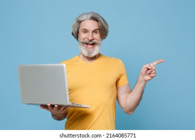 Fun Elderly Gray-haired Mustache Bearded Man 50s In Casual Yellow T-shirt Hold Laptop Pc Computer Pointing Index Finger Aside Isolated On Blue Background. People Lifestyle Concept. Mock Up Copy Space 