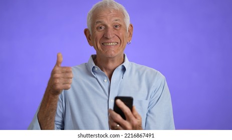 Fun Excited Surprised Elderly Gray-haired Man 70s In Blue Shirt Hold Mobile Phone And Smile Give Thumbs Up, Celebrate Success, Isolated On Solid Purple Background Studio Portrait 