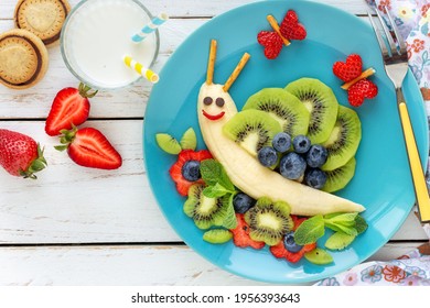 Fun food for kids - cute smiling snail made of fresh fruits (bananas, kiwi, blueberries and strawberries) as a healthy breakfast for children served with milk and cookies