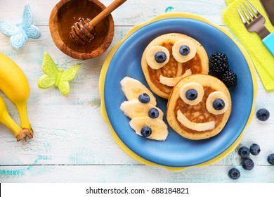 Fun food for kids - cute smiling faces on sweet pancakes for breakfast with bananas, fresh blueberries and blackberries. Creative cooking for children