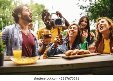 Fun and Friendship at Outdoor Bar Counter. Diverse group of friends enjoying drinks, singing, and dancing to music played by young man with dreadlocks. Scenic backdrop of trees. - Powered by Shutterstock