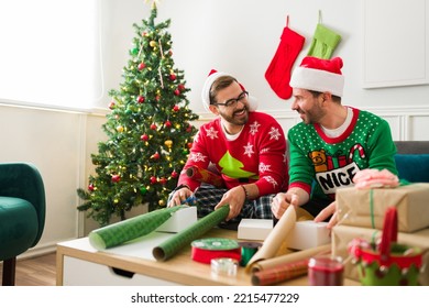 Fun Gay Couple In Pajamas Laughing While Wrapping Christmas Presents Together For The Holidays In The Living Room